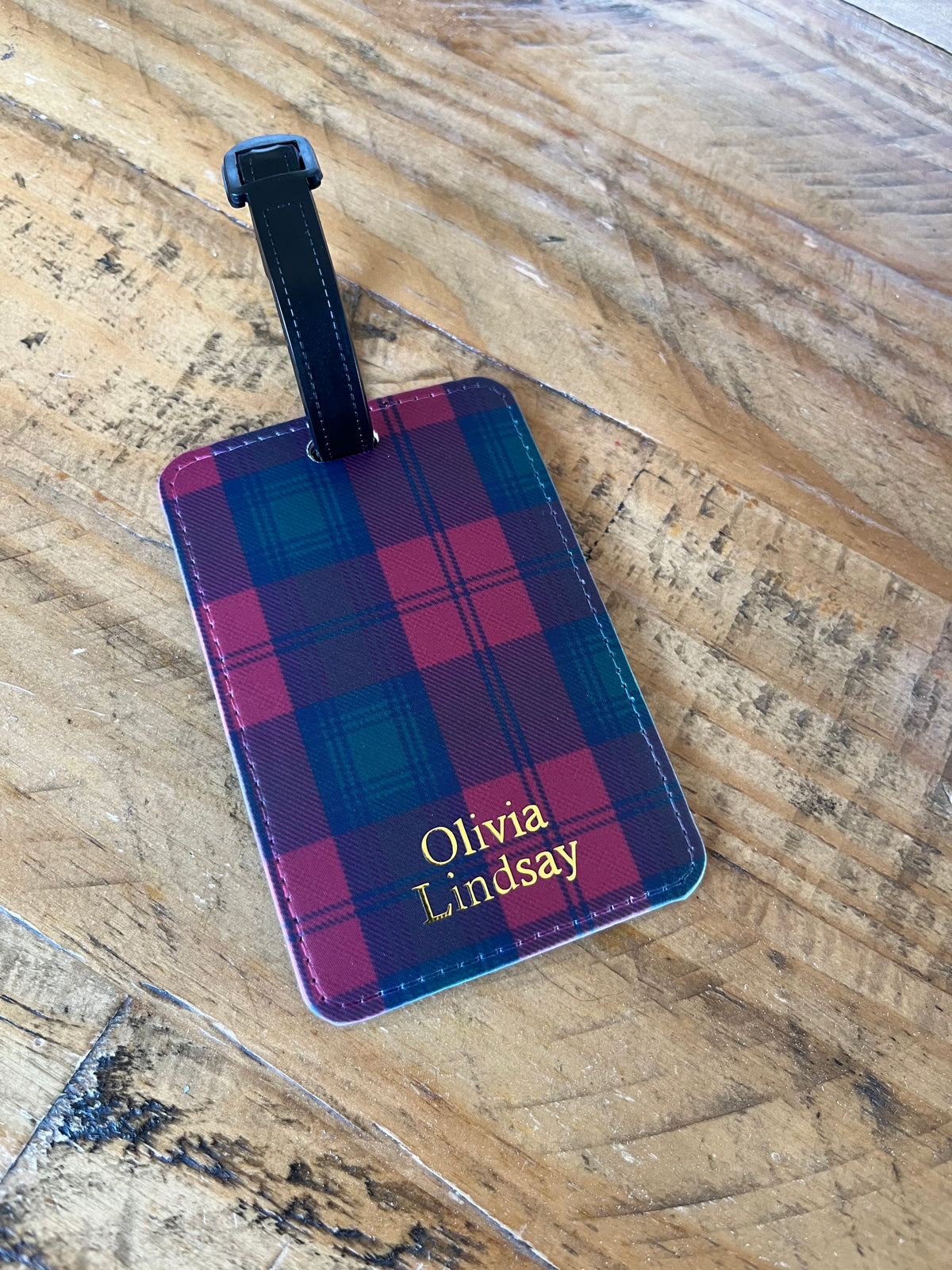 A rectangular luggage tag with a red and green plaid pattern, personalized with gold HTV reading 'Olivia Lindsay', resting on a rustic wooden surface.
