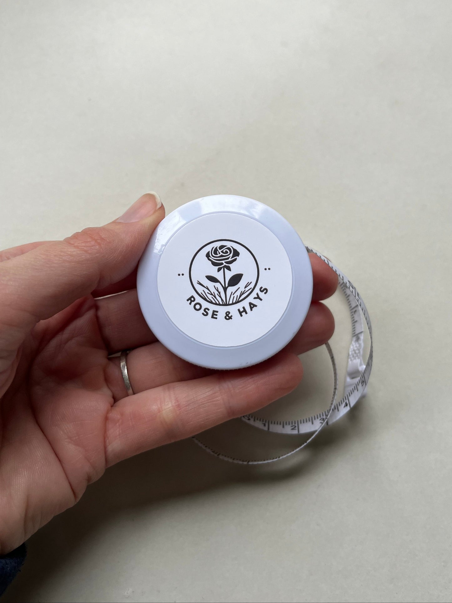 Hand holding a circular tape measure with 'Rose & Hays' logo in black on a plain background