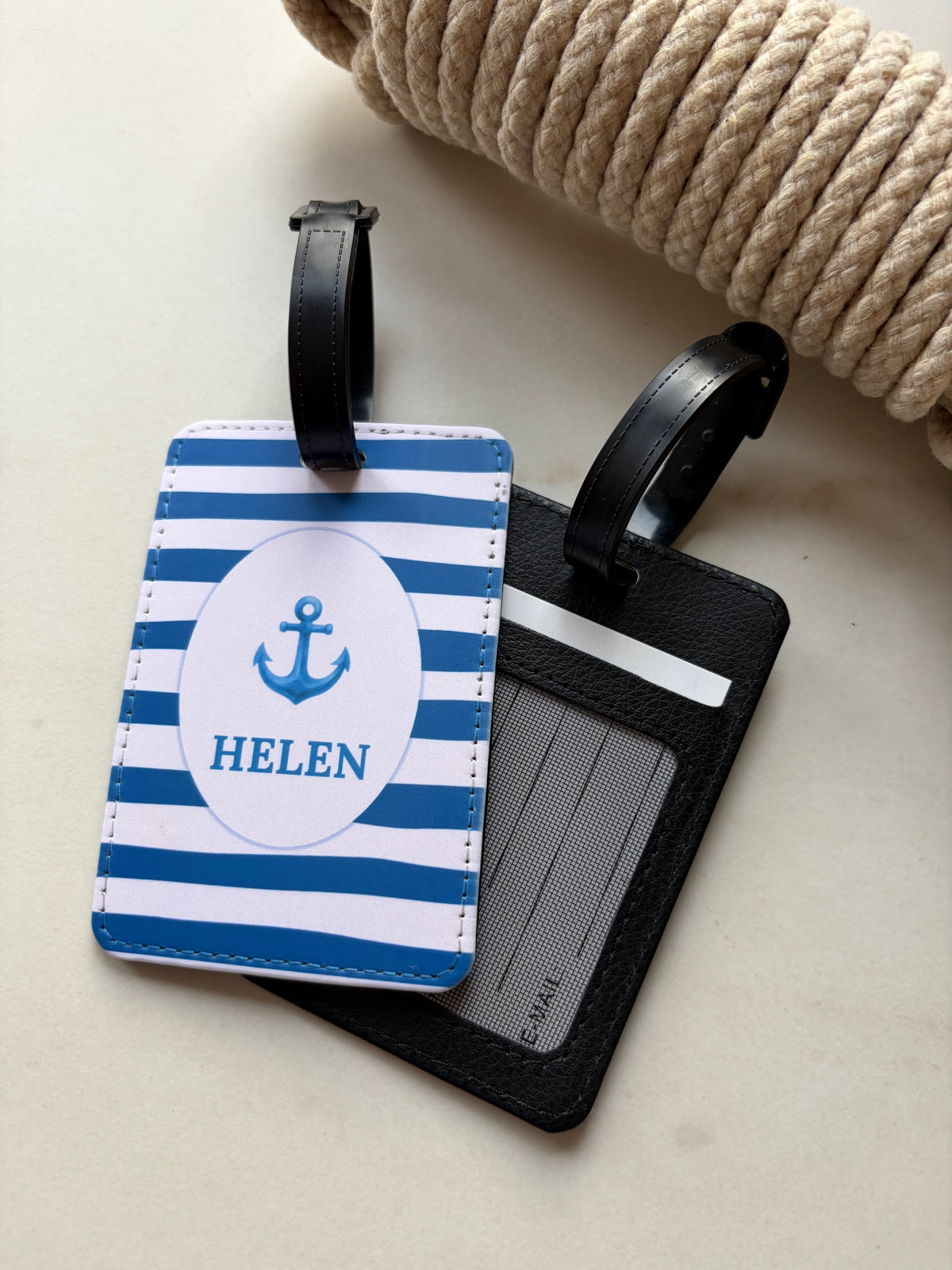 A personalized nautical-themed luggage tag set on a light beige surface with a rolled rope prop. One tag features blue and white stripes with a white oval containing a blue anchor and the name 'HELEN'.
