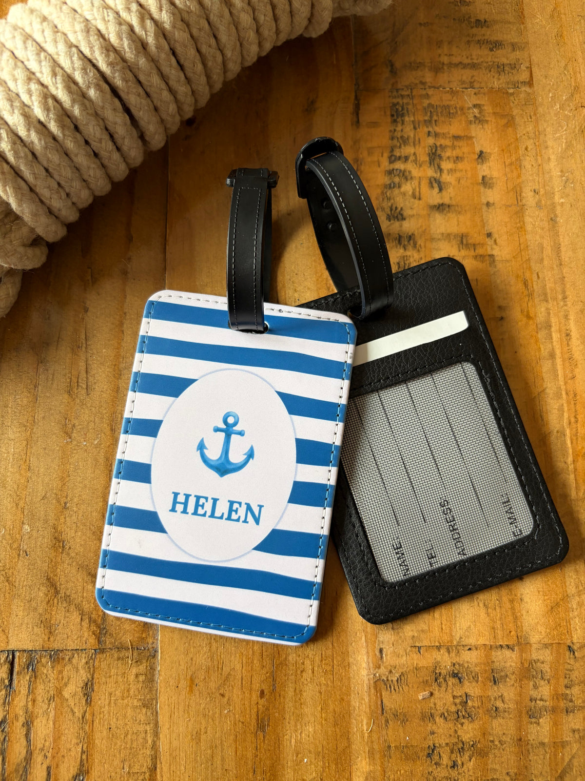 Personalized nautical-themed luggage tags on a rustic wooden surface with a coiled rope prop. One tag features blue and white stripes with a blue anchor and the name 'HELEN' in a white oval.