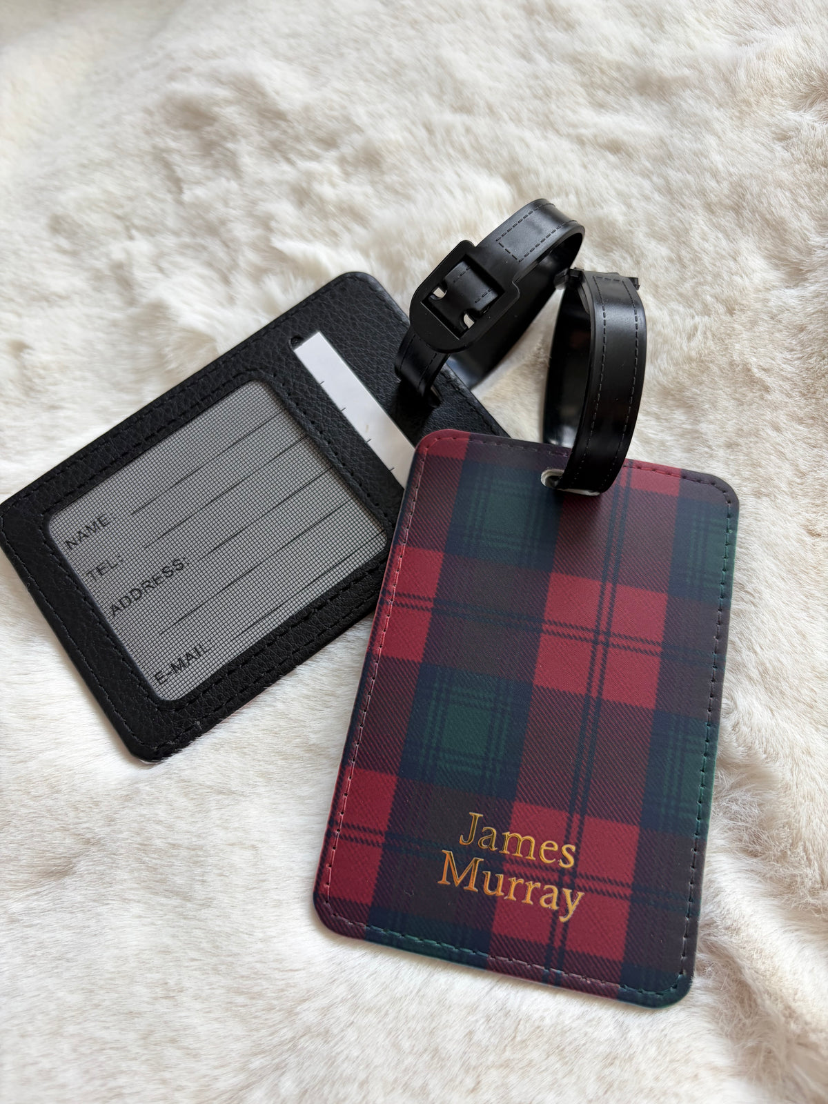 Personalized luggage tag set on a white faux fur background. One tag is black with a clear ID window and black strap, and the other is a red and green tartan plaid tag with gold text reading 'James Murray' and a black strap.