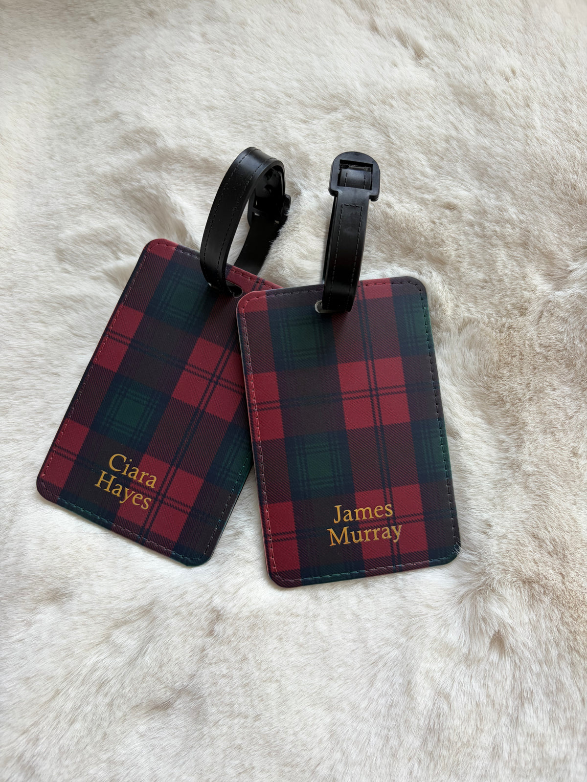 Two personalized luggage tags with a red and green tartan plaid pattern and black straps, lying on a white faux fur surface. One tag is personalized with 'Ciara Hayes' and the other with 'James Murray' in gold lettering.
