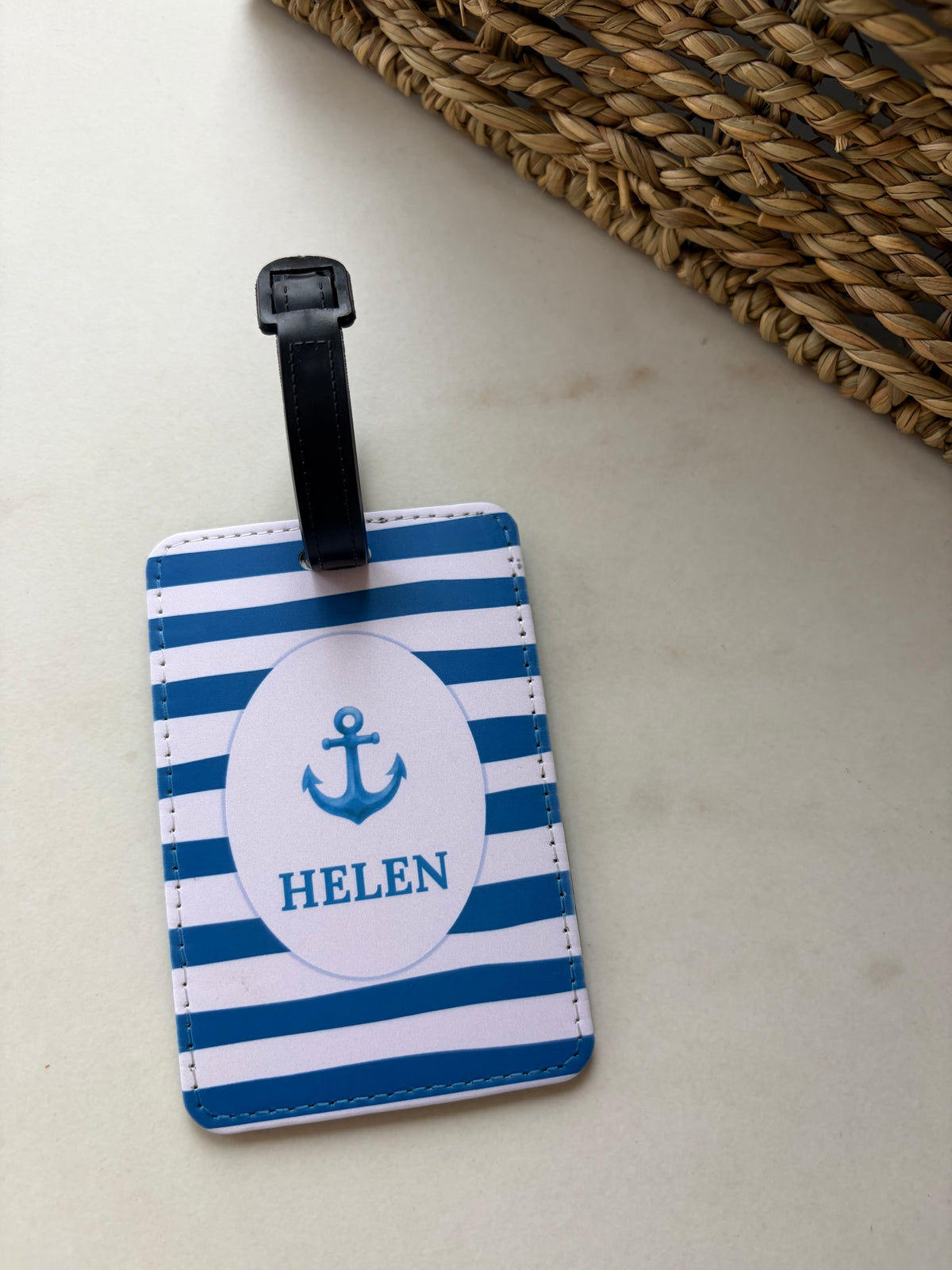 A personalized luggage tag with blue and white horizontal stripes, a black strap, and a white oval center featuring a blue anchor and the name 'HELEN'.
