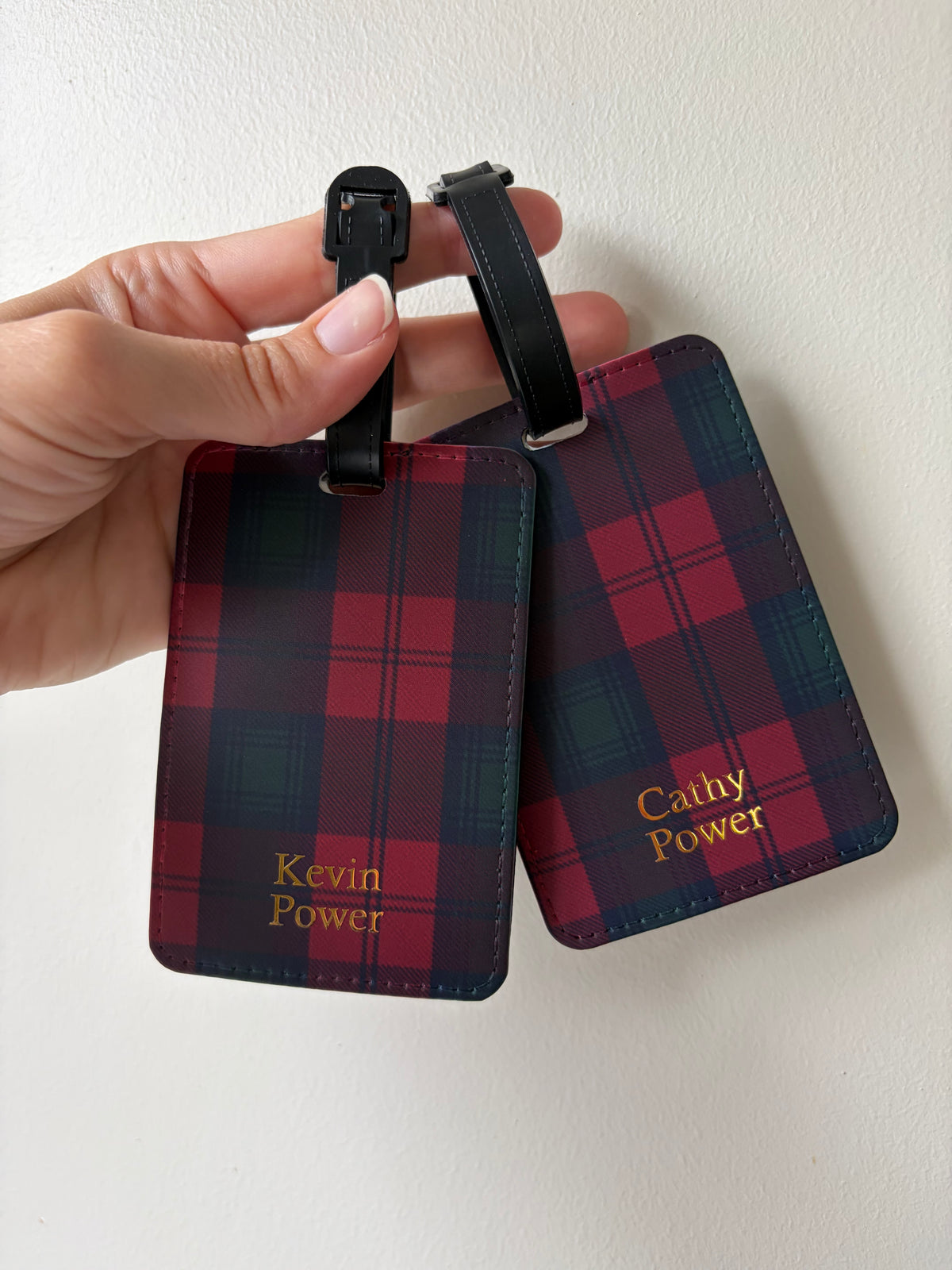A hand holds two personalized luggage tags with a red and dark green tartan plaid pattern and black straps against a plain off-white wall. The tags are personalized with the names 'Kevin Power' and 'Cathy Power' in gold foil.