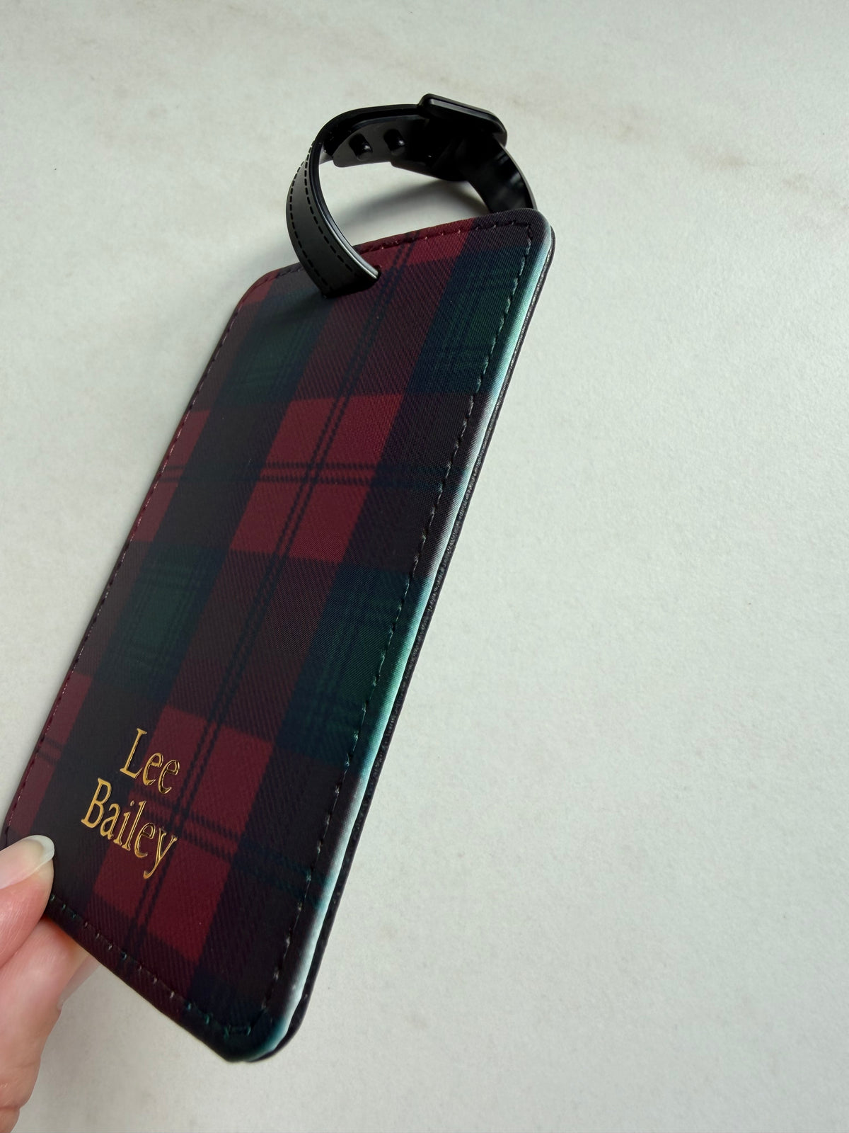 A personalized luggage tag with a red and green tartan plaid pattern, featuring gold embroidered text reading 'Lee Bailey' and a black strap, held by a hand against a light gray background.