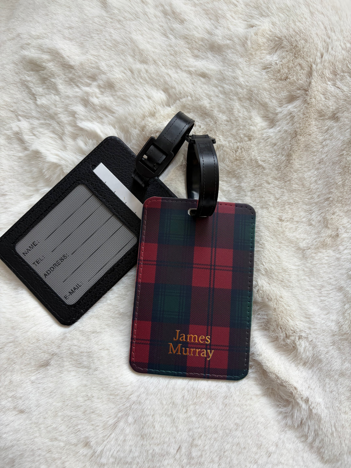 A personalized luggage tag set featuring a red and green tartan plaid name tag with gold 'James Murray' text, and a black ID window holder, displayed on a white faux fur background.