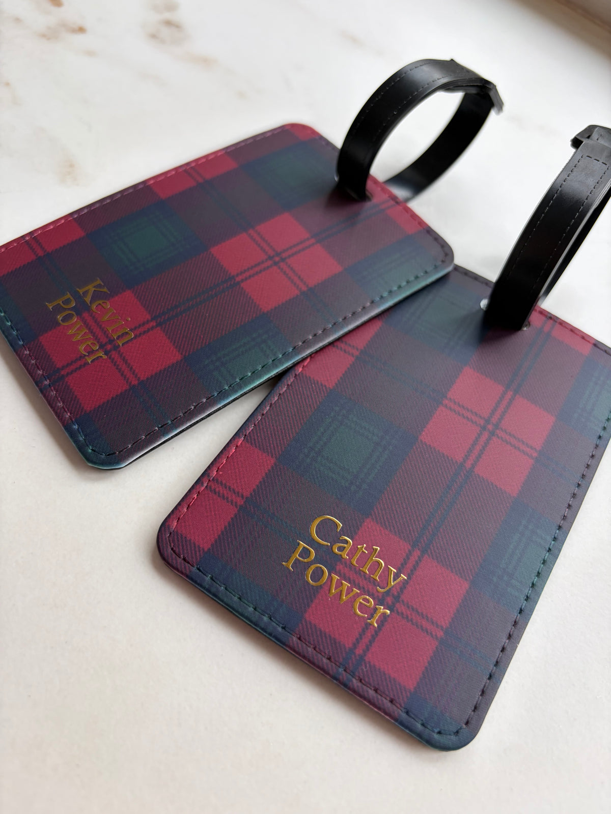 Two personalized luggage tags with a red and navy tartan plaid pattern and black straps, featuring the name 'Cathy Power' in gold foil, displayed on a light marble surface.