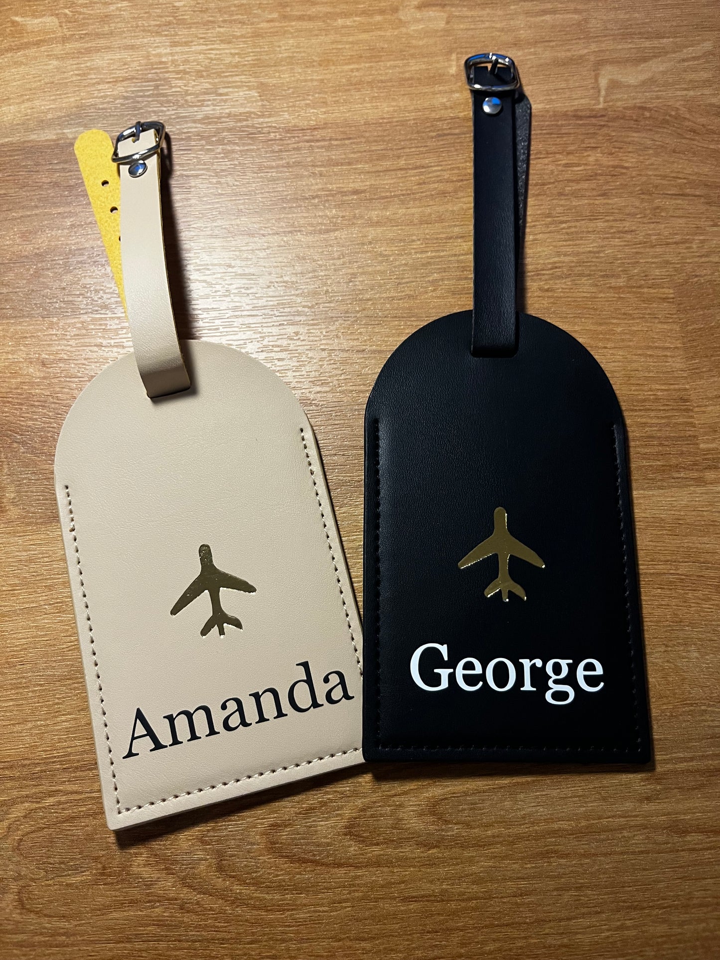 Two luggage tags, one black with 'George' and one beige with 'Amanda', on a wooden surface.