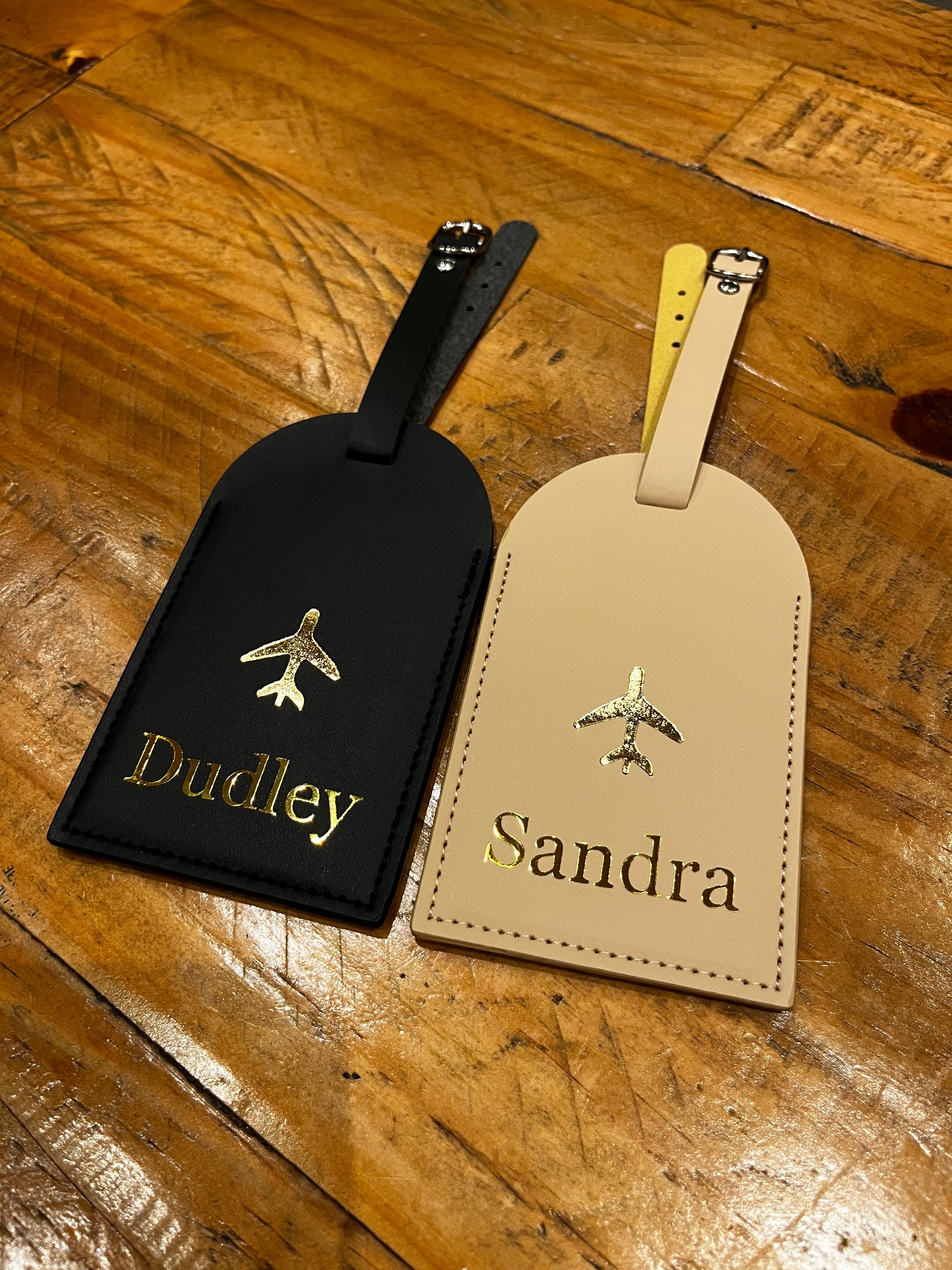Two luggage tags, one beige and one black, with 'Sandra' and 'Dudley' personalised luggage tags on a wooden surface.