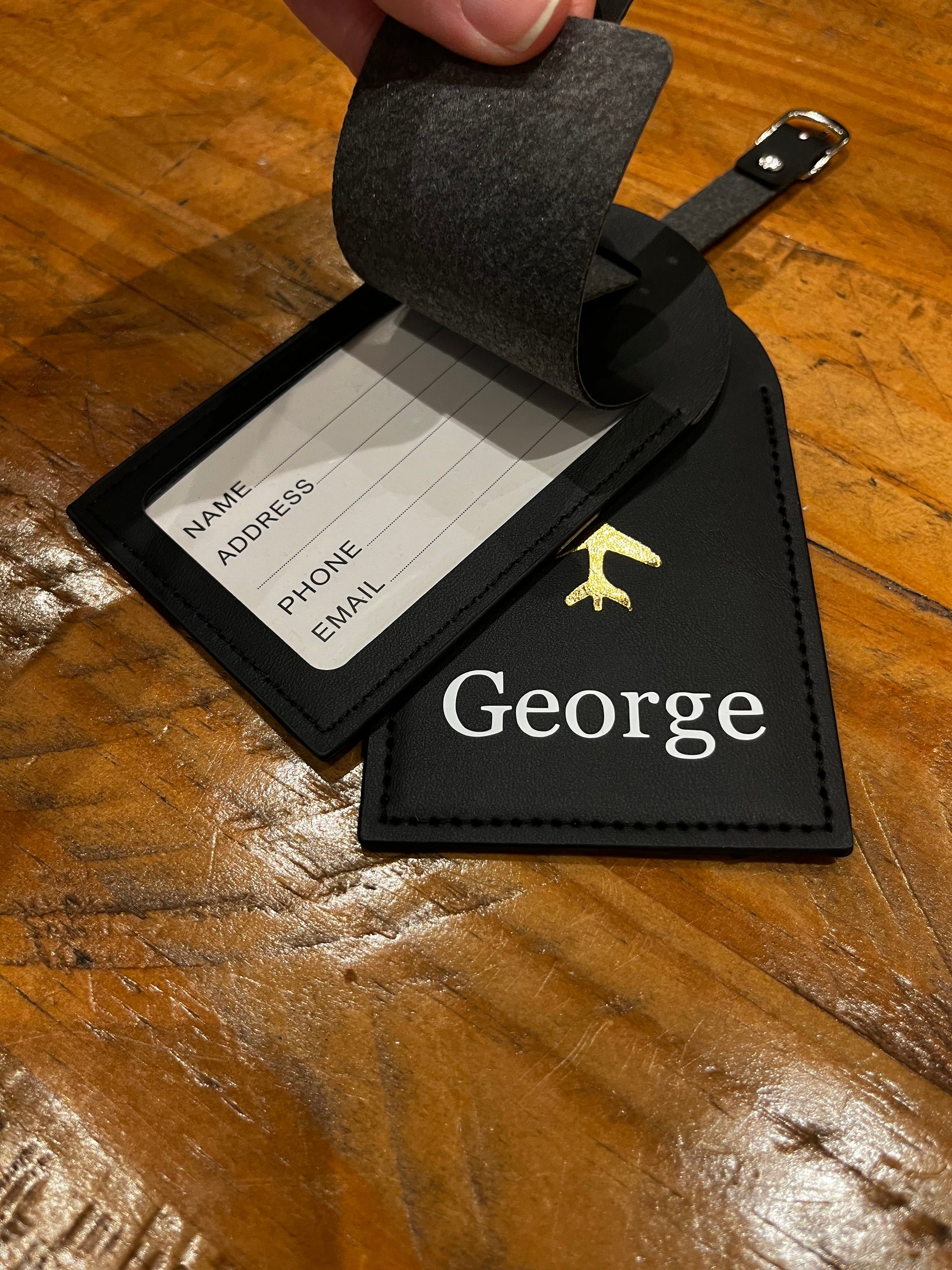 Black luggage tag with 'George' added in white on a wooden surface